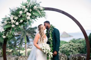 A bride and groom kissing in front of a wedding arched decorated by white roses with the ocean and chinaman's hat in the background