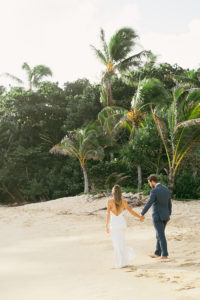 Bride and Groom walking on a sandy beach full of tropical trees captured by Rachel Kathryn