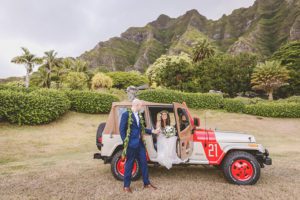 Groom assisting bride to get off a jeep in front of tropical trees and mountains in Kualoa Valley captured by Vivid Fotos