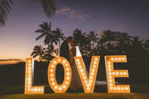 A bride and groom in front of a LOVE during Sunset Photoshoot with Coconut trees in the background captured by Vivid Fotos