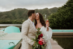 A wedding couple kissing in front of Kualoa Mountains while the bride holds her Gazoz cocktail