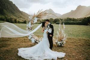 Bride and groom share a kiss in front of a beautifully decorated wedding arch, while the bride's veil gently flutters in the air at Kualoa Ranch captured by Seeking Films