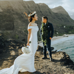 Bride and Groom standing on a cliff overlooking the ocean against tropical mountains in Hawaii by Seeking Films