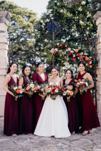 A bride and her bridesmaids holding a bouquet of tropical flowers in front of a gate decorated by flowers