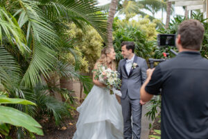 Bride and Groom smiling at each other at a tropical garden while being filmed by Aloha Films