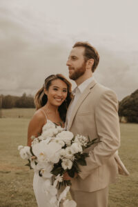 A bride, showcasing her hair and make up done by Perfectly Made Hawaii while holding a big bouquet of white flowers, and her groom on their wedding day