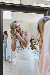 A Blonde bride wearing a veil and wedding dress getting ready in front of a mirror on her wedding day